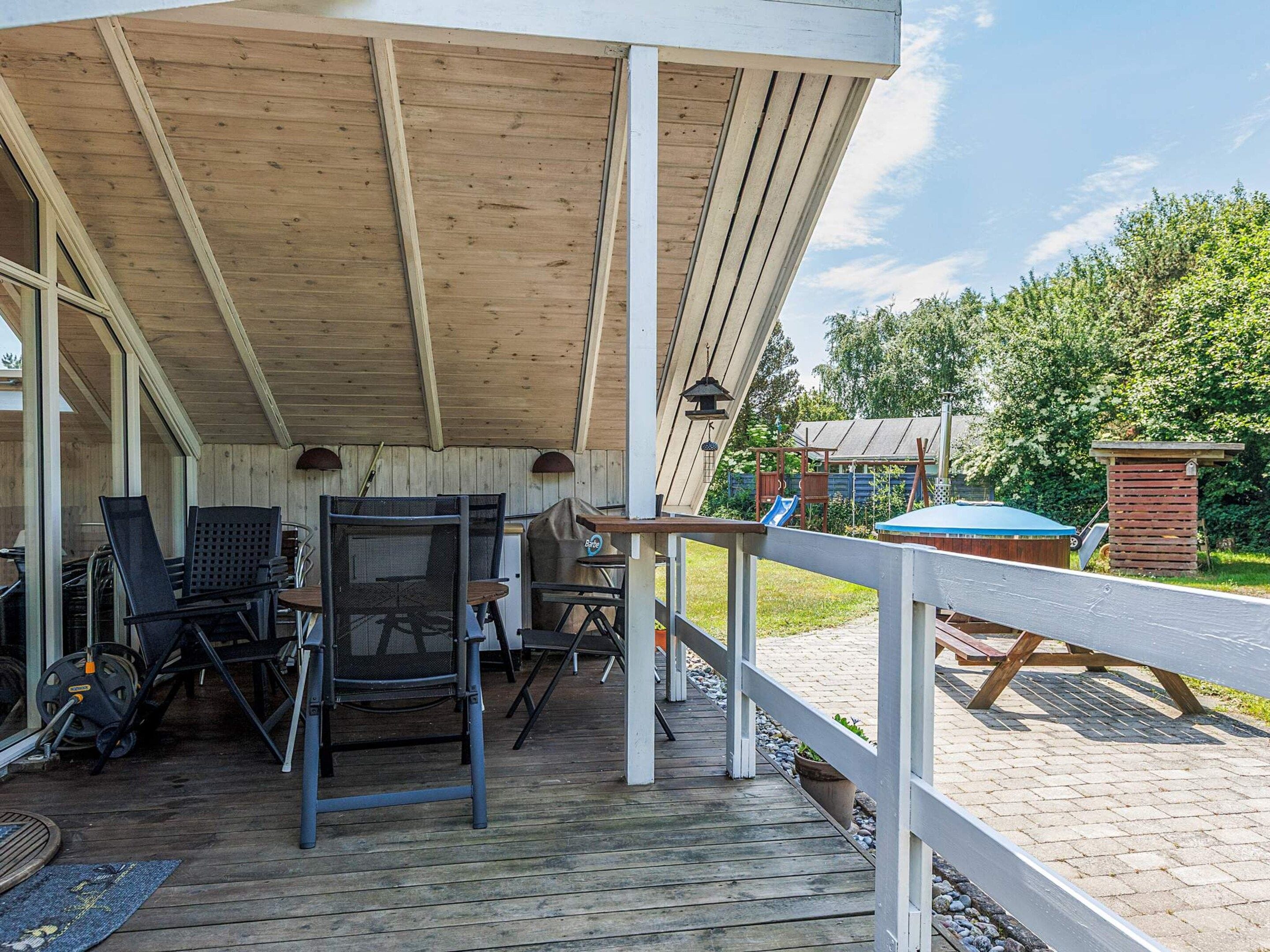 A covered outdoor patio with a wooden deck, metal chairs, a barbecue grill, and a small table.