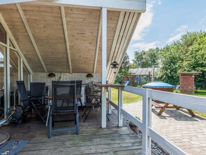 A covered outdoor patio with a wooden deck, metal chairs, a barbecue grill, and a small table. - 5 Star Holiday Home in Dannemare (Dannemare)