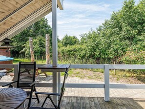A covered outdoor seating area with tables and chairs, a wooden deck, and a view of a wooded area.