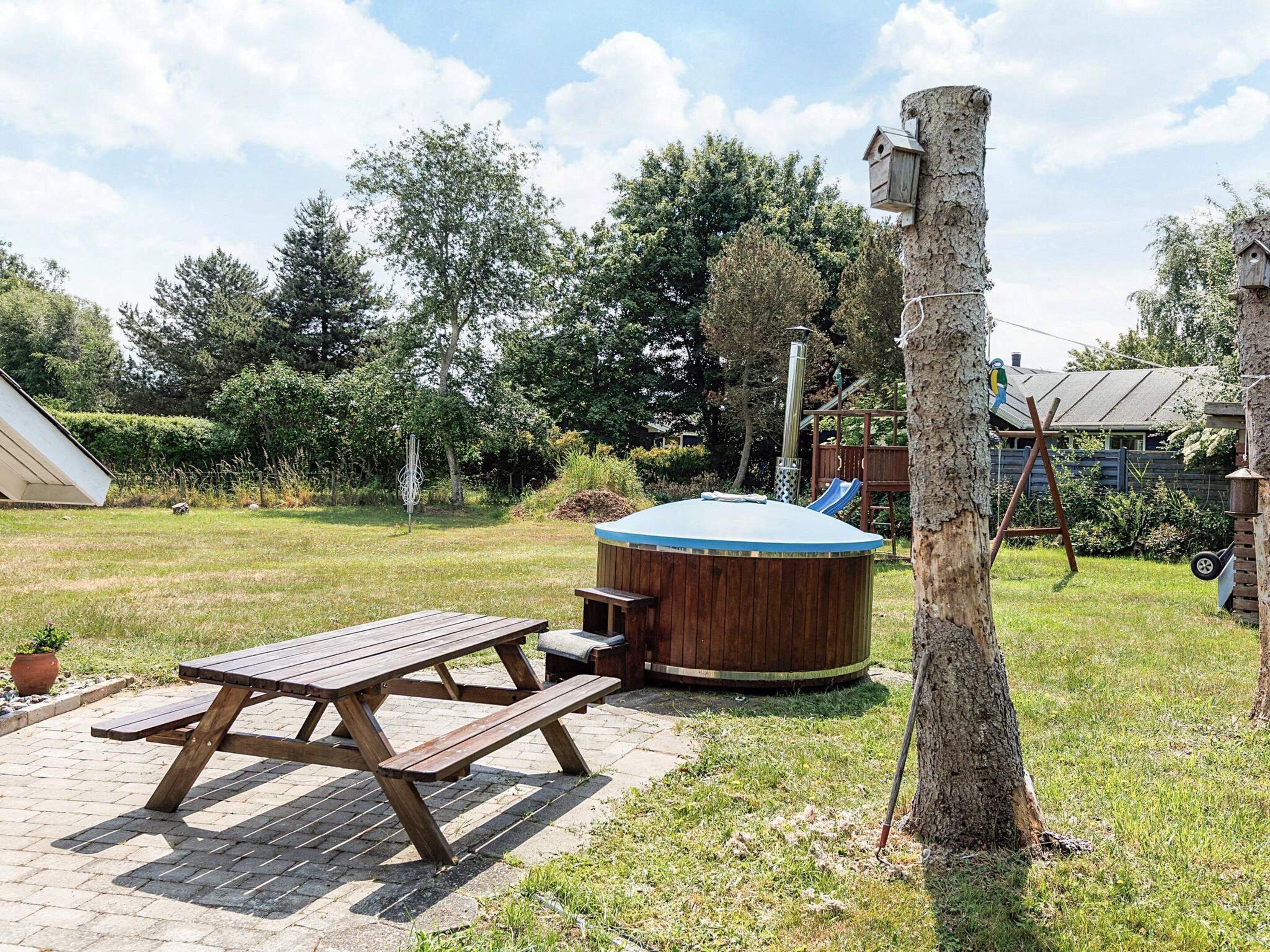 A wooden picnic table and hot tub in a backyard with trees and a play structure in the background.