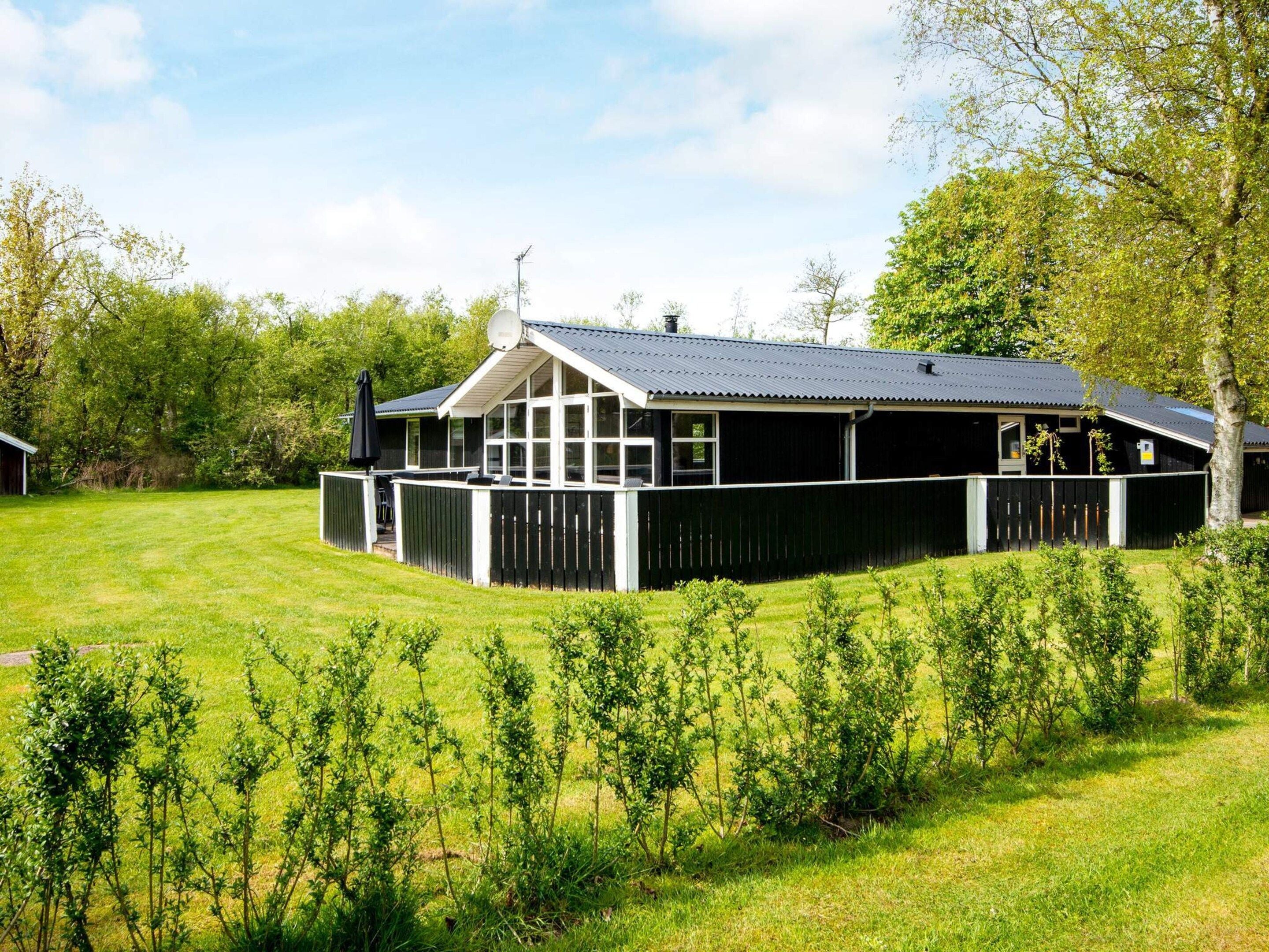 Ein eingeschossiges Gebäude mit dunklem Dach und weißer Fassung, umgeben von einem eingezäunten Garten und Bäumen.