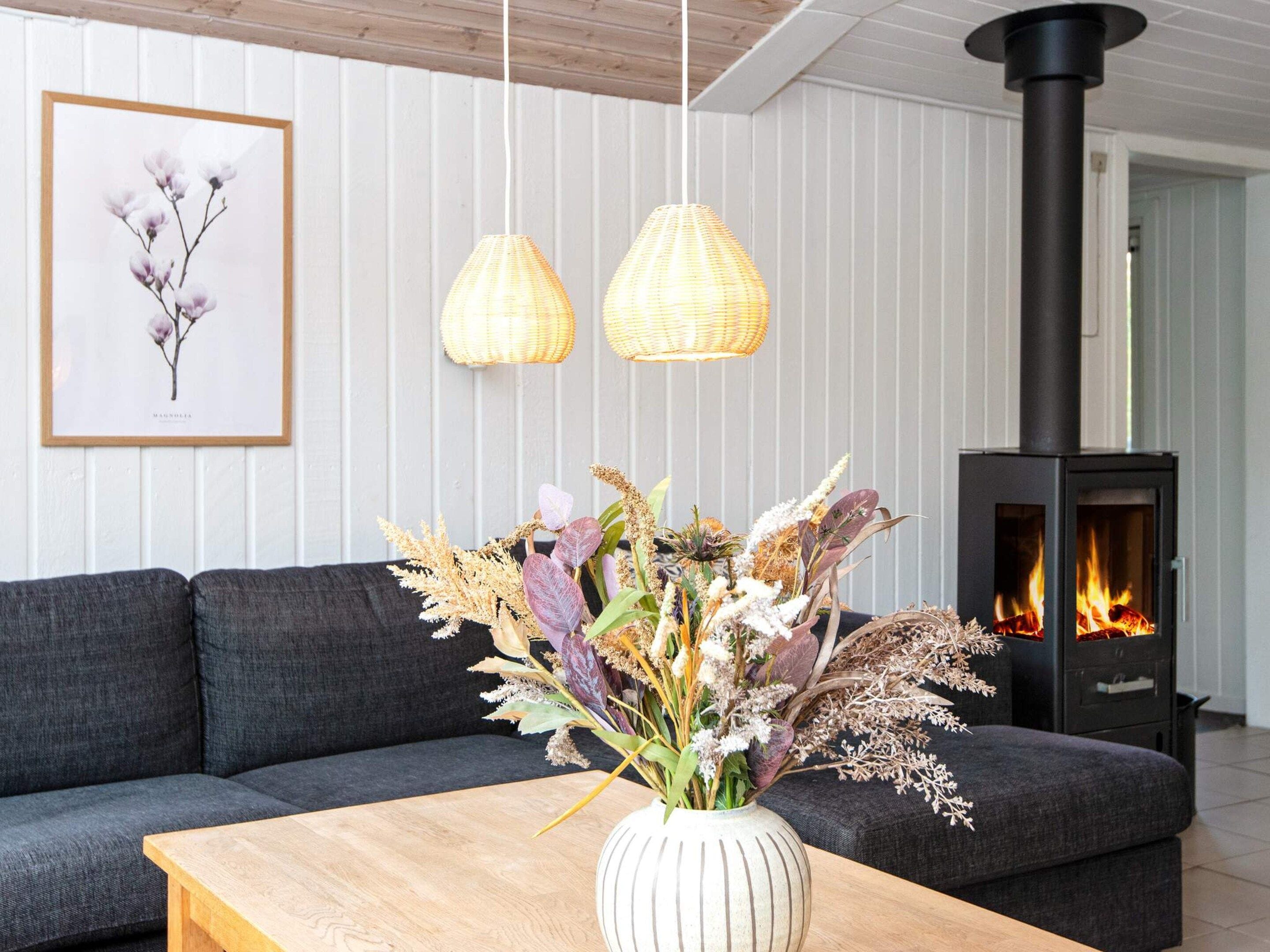 A modern living room with a sofa, a wooden coffee table, a vase with dried flowers, two hanging lamps, and a wood-burning stove.