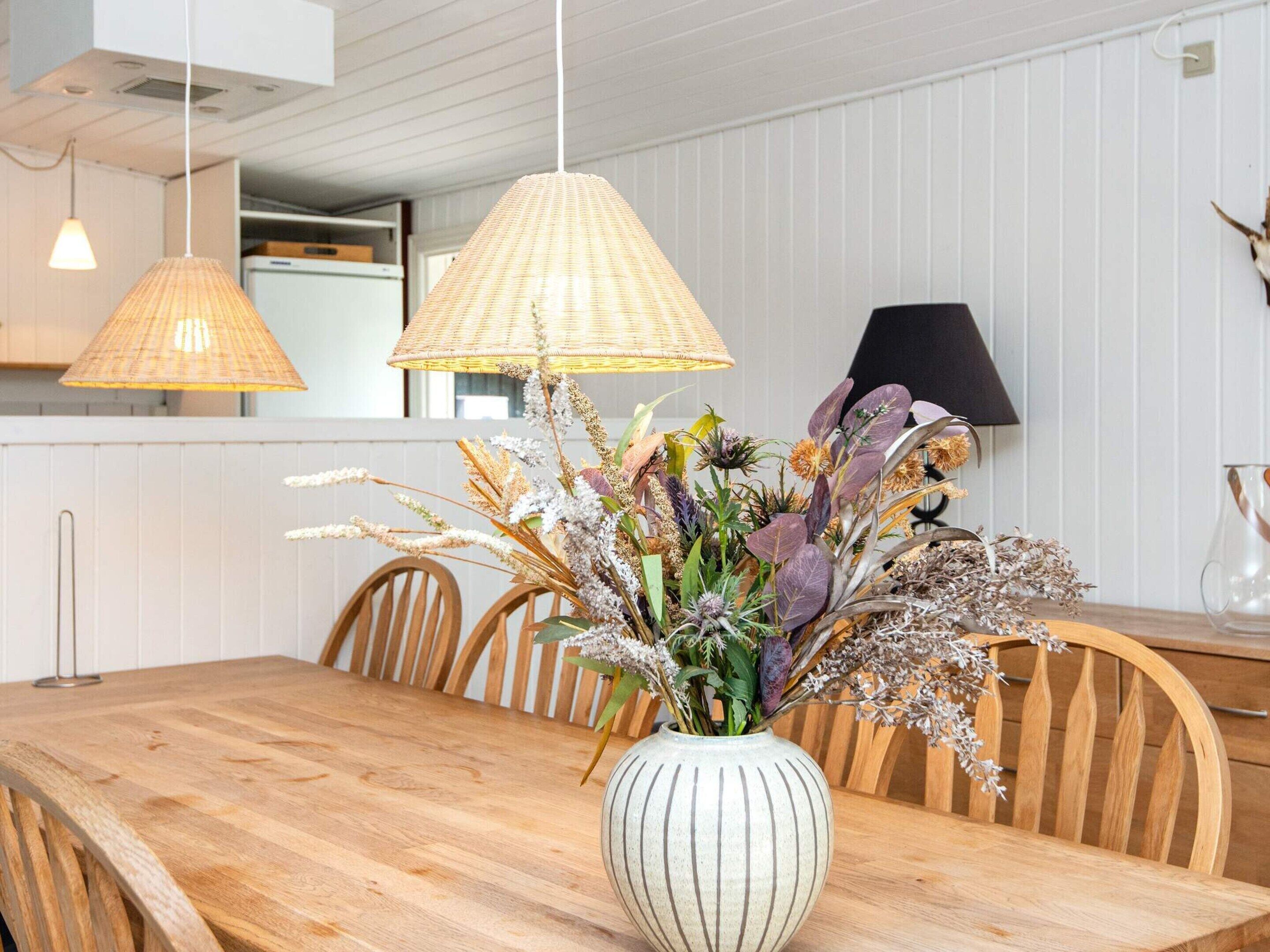 A dining area with a wooden table, two hanging lamps, and a vase with dried flowers.