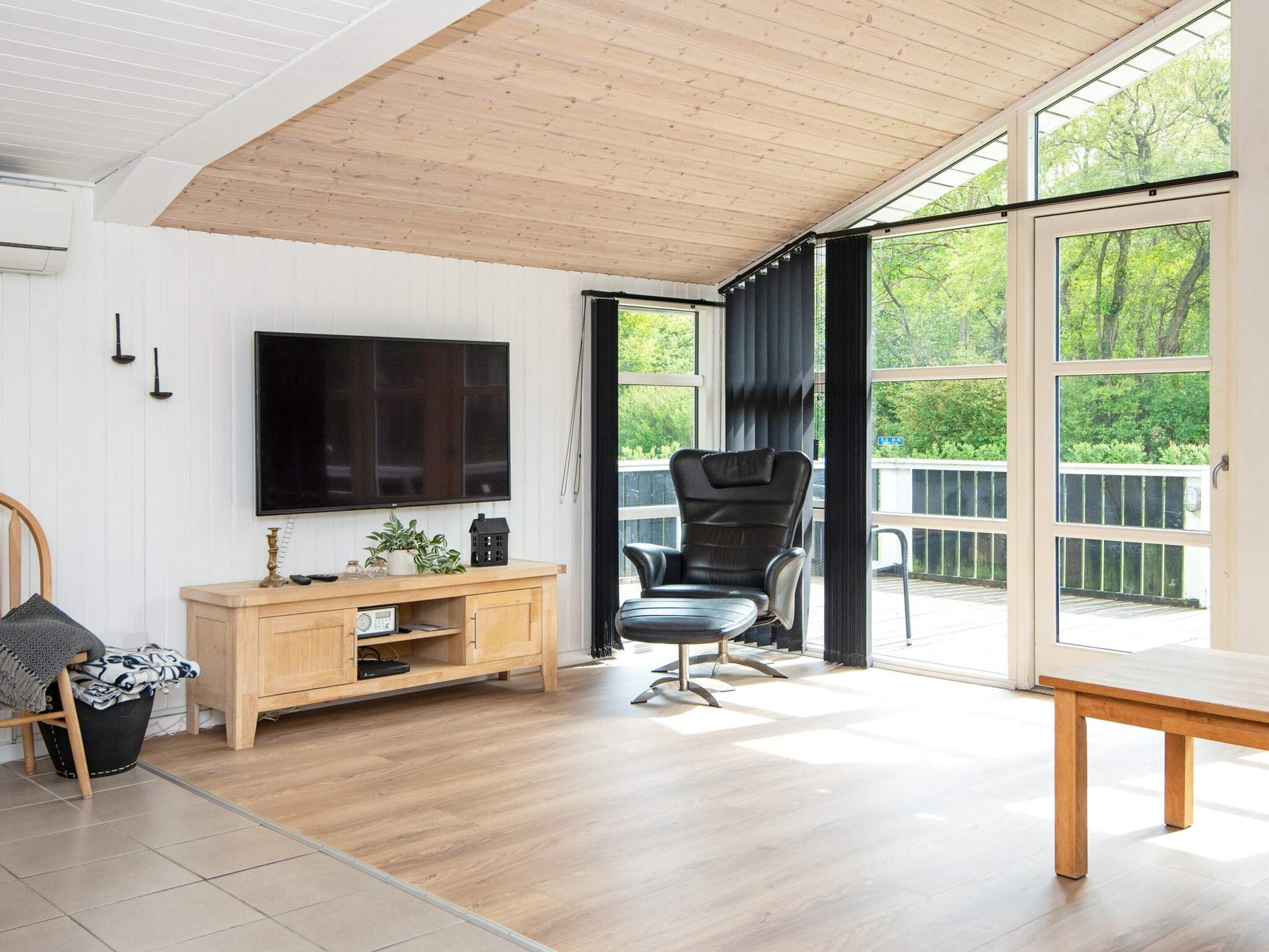 Un salon moderne avec un plafond en bois, un téléviseur à écran plat, un meuble en bois, un fauteuil en cuir noir et une grande fenêtre donnant sur un paysage verdoyant.
