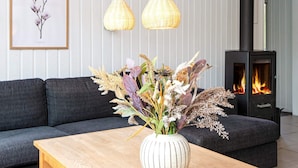 Ett modernt vardagsrum med en soffa, ett soffbord i trä, en vas med torkade blommor, två hängande lampor och en vedkamin.