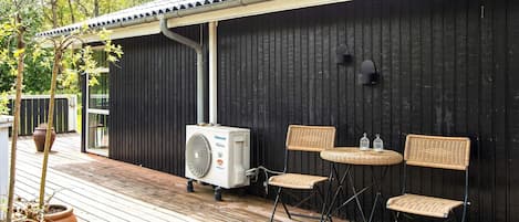 A covered outdoor area with a wooden deck, two wicker chairs, a small table, and a portable air conditioning unit.