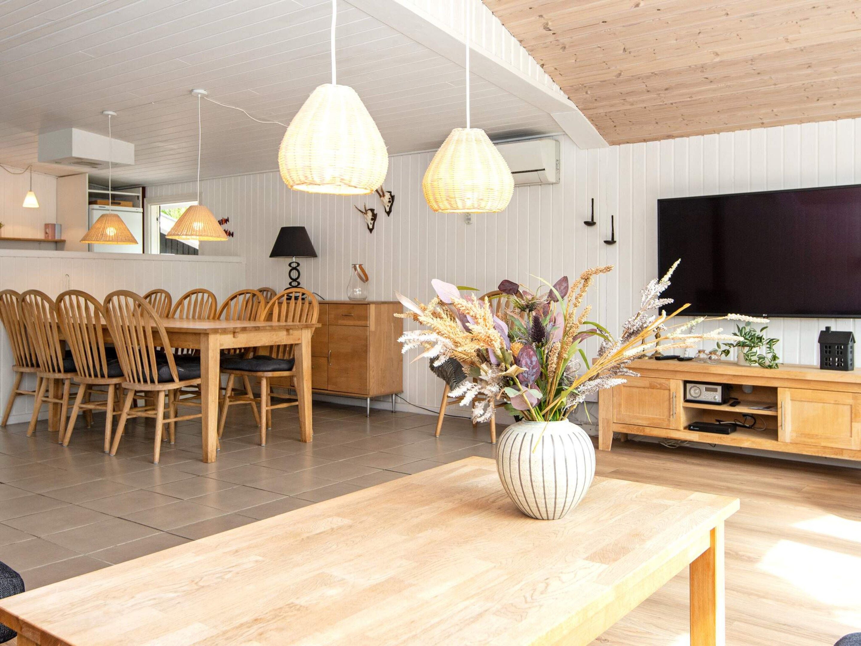 A modern living room with a wooden dining table, chairs, a large TV, and a decorative vase.