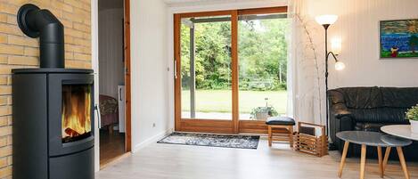 A modern living room with a wood-burning stove, a sofa, a coffee table, and a view of a garden through a glass door.