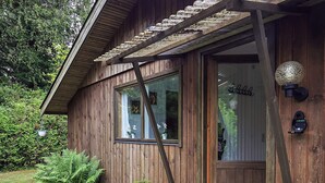 Ein Holzhaus mit Veranda, einer Tür mit Glasfenster und einem kleinen Garten mit Farne und einer Nadelbaumart.