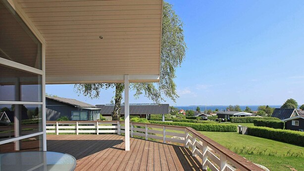 Une terrasse en bois avec une table en verre, des rampes blanches et une vue sur la mer et les maisons au loin.