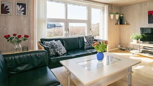 A modern living room with a black leather sofa, a glass coffee table, and a television. There are windows with white curtains, a wooden ceiling, and a plant on a shelf. - 4 Star Holiday Home in Lokken (Lokken)