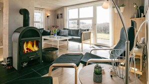 A modern living room with a wood-burning stove, comfortable seating, and large windows offering a view of the outdoors. - 4 Star Holiday Home in Lokken (Lokken)