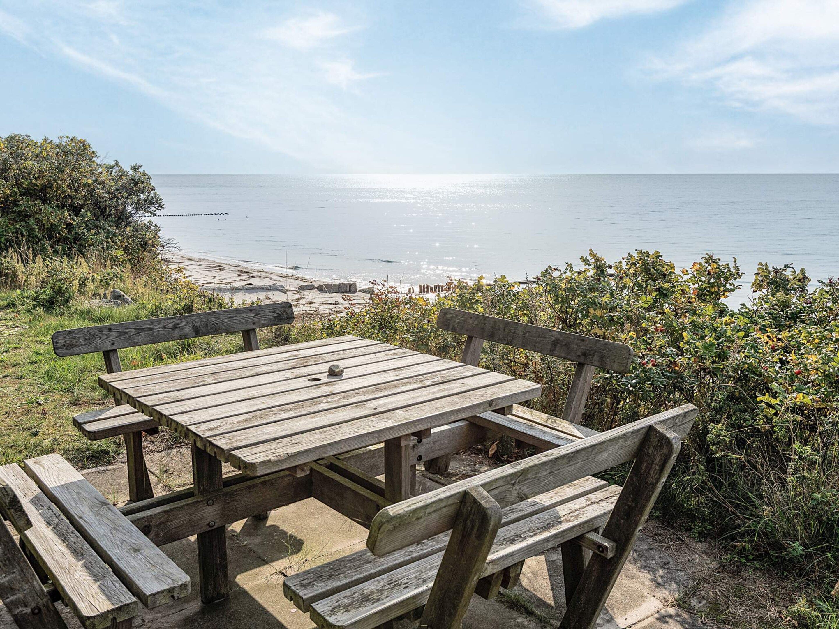 A wooden picnic table with benches, overlooking a beach with calm waters and distant shoreline.