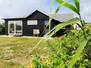 A modern black house with a glass door entrance, a small patio with a table and chairs, and a garden with green plants and a pink flower. - 4 Star Holiday Home in Ringkobing (Ringkobing)