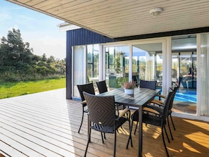 An outdoor deck with a table and chairs, a glass door leading to a pool, and a view of a garden. - 5 Star Holiday Home in Ringkobing (Ringkobing)