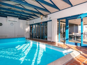 An indoor swimming pool with clear water, a ceiling with blue beams, and a view of the outside patio area. - 5 Star Holiday Home in Ringkobing (Ringkobing)