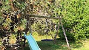 A wooden swing set with a green slide in a grassy area with trees. - 7 Person Holiday Home in Blavand (Blavand)