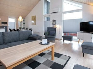 Una sala de estar moderna con sofá, sillón y reposapiés, un comedor con sillas, un televisor y un ventanal grande.