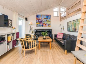 A living room with a wooden floor, a black leather sofa, a wooden coffee table, and a black leather couch. There is a painting on the wall, a TV on the left, and a bookshelf with books and decorative items.