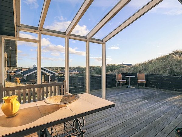A wooden deck with a glass enclosure, a table with chairs, and a view of the surrounding landscape.