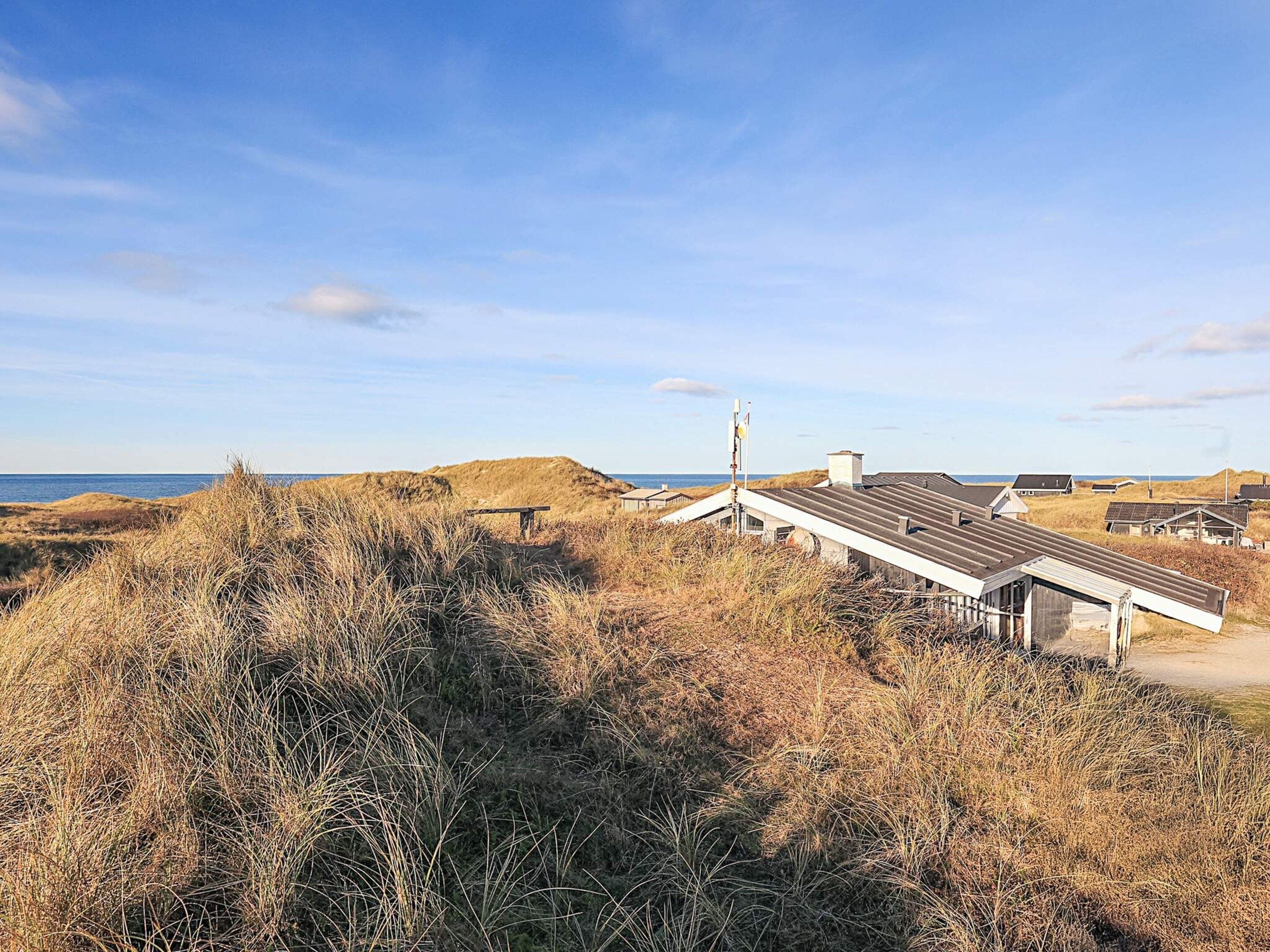 A coastal building with a flat roof, surrounded by dry grass and dunes, with the sea in the background.