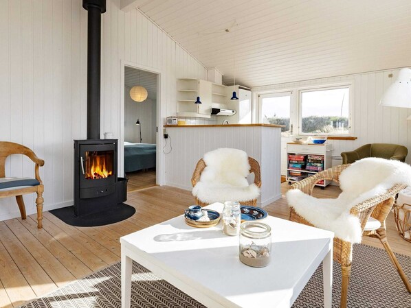 Une pièce chaleureuse avec un poêle à bois, des fauteuils en osier, une table blanche ornée de vases décoratifs et une vue sur l’extérieur par une fenêtre.