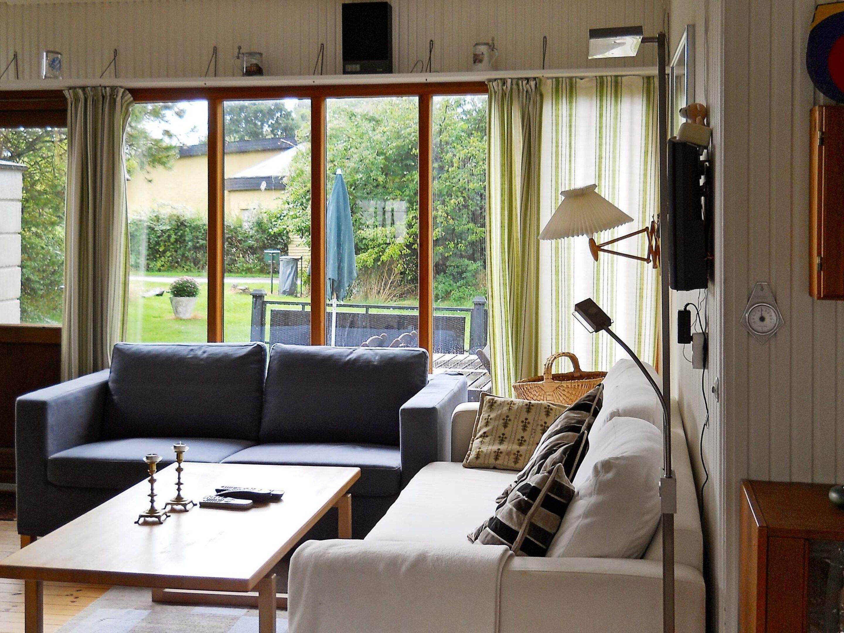 En stue med sofa, sofabord og lampe. Store vinduer med gardiner, udsigt til det fri og et væghængt ur.