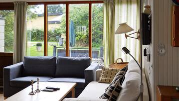A living room with a sofa, a coffee table, and a lamp. There are large windows with curtains, a view of the outdoors, and a wall-mounted clock.