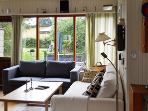 A living room with a sofa, a coffee table, and a lamp. There are large windows with curtains, a view of the outdoors, and a wall-mounted clock.