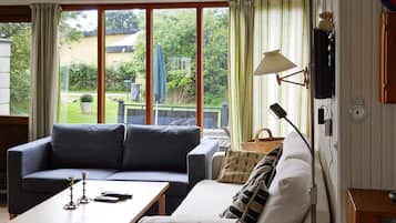 A living room with a sofa, a coffee table, and a lamp. There are large windows with curtains, and a view of the outdoors.