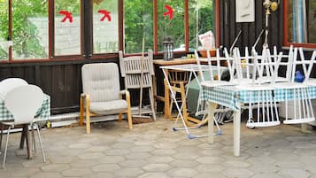 A covered outdoor seating area with hexagonal tiles, wooden chairs, a table with a checkered tablecloth, and a view of greenery through large windows.