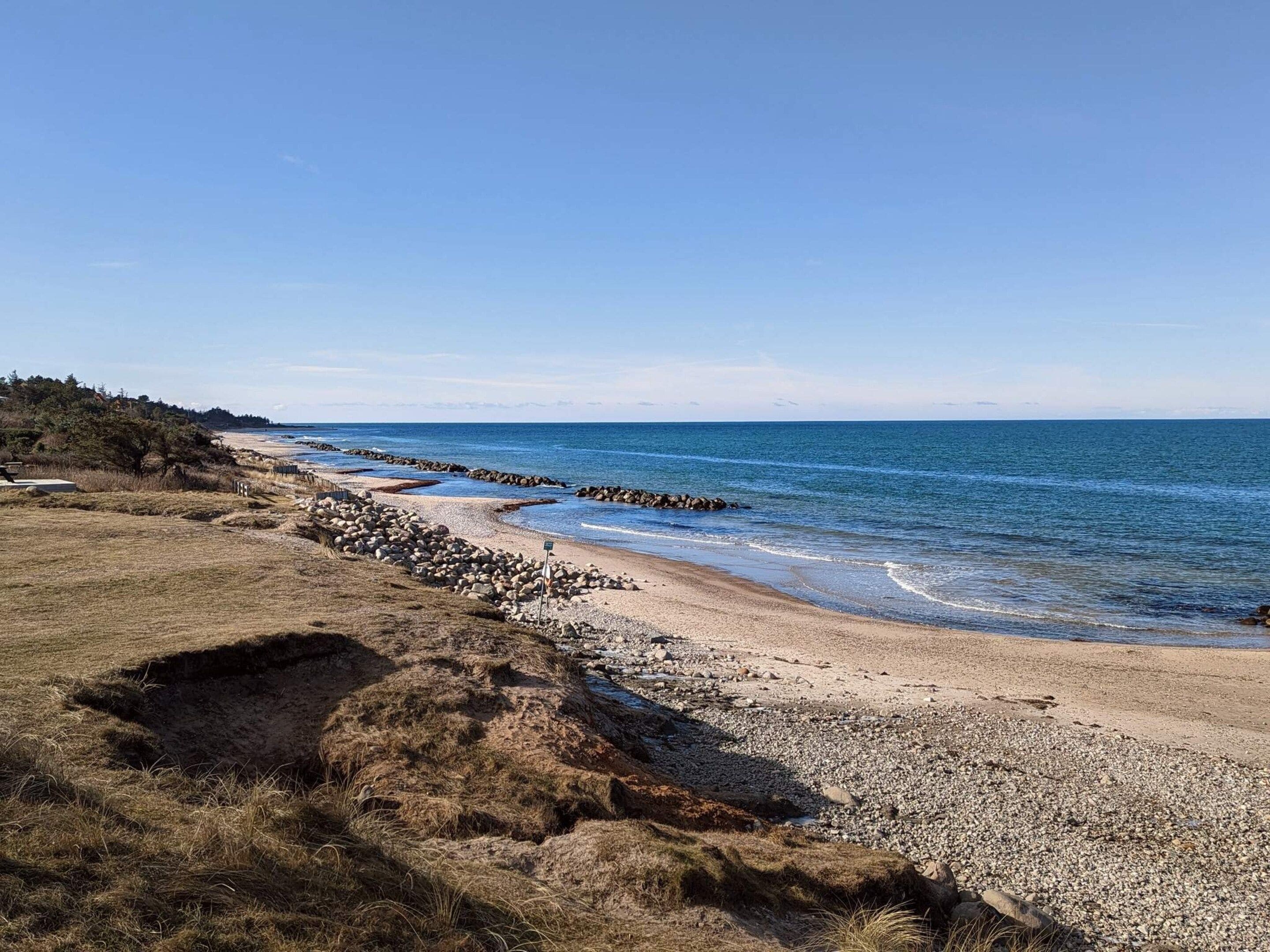 Et kystlandskab med en stenstrand, en klippefyldt bølgebryder og en klar himmel.