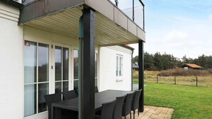 A covered outdoor patio with a table and chairs, a white building with large windows, and a grassy area in the background. - 8 Person Holiday Home in Lokken-by Traum (Lokken)