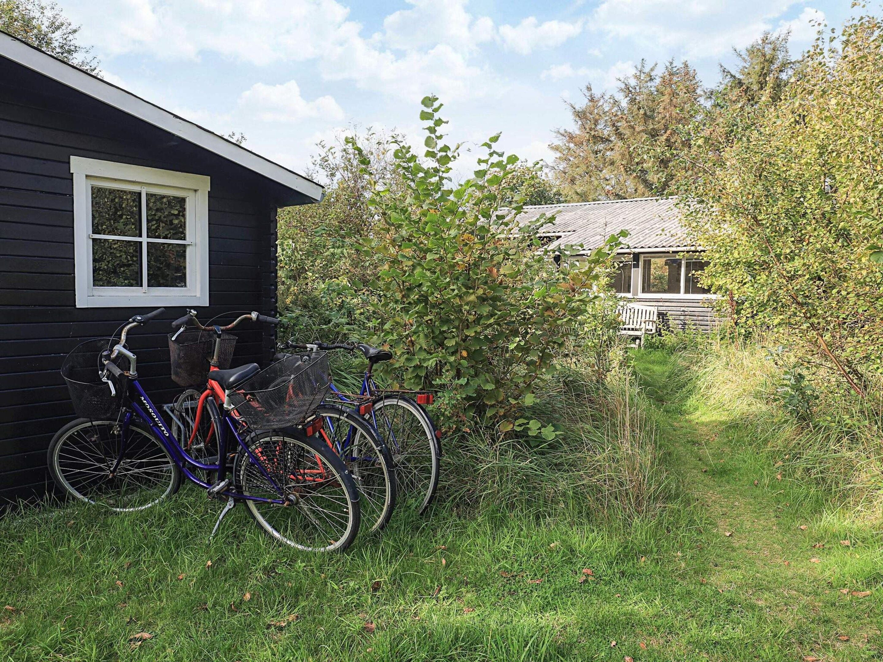 Een zwart gebouw met een raam, een fiets ervoor geparkeerd, en een pad dat naar een ander gebouw met een veranda leidt.