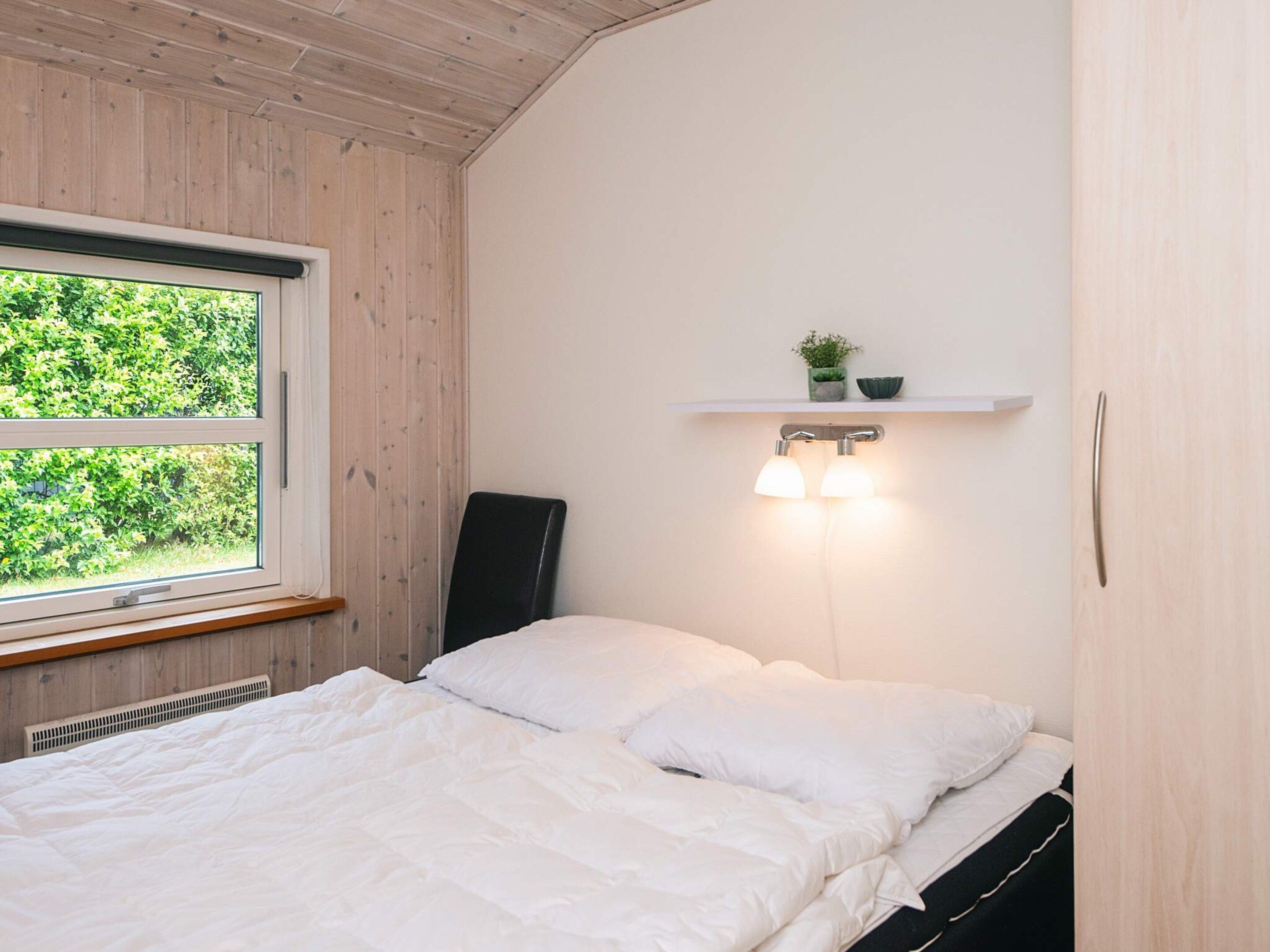 Et soveværelse med seng, et vindue med udsigt over grøn natur, en vægmonteret hylde med en plante og en vægmonteret lampe.
