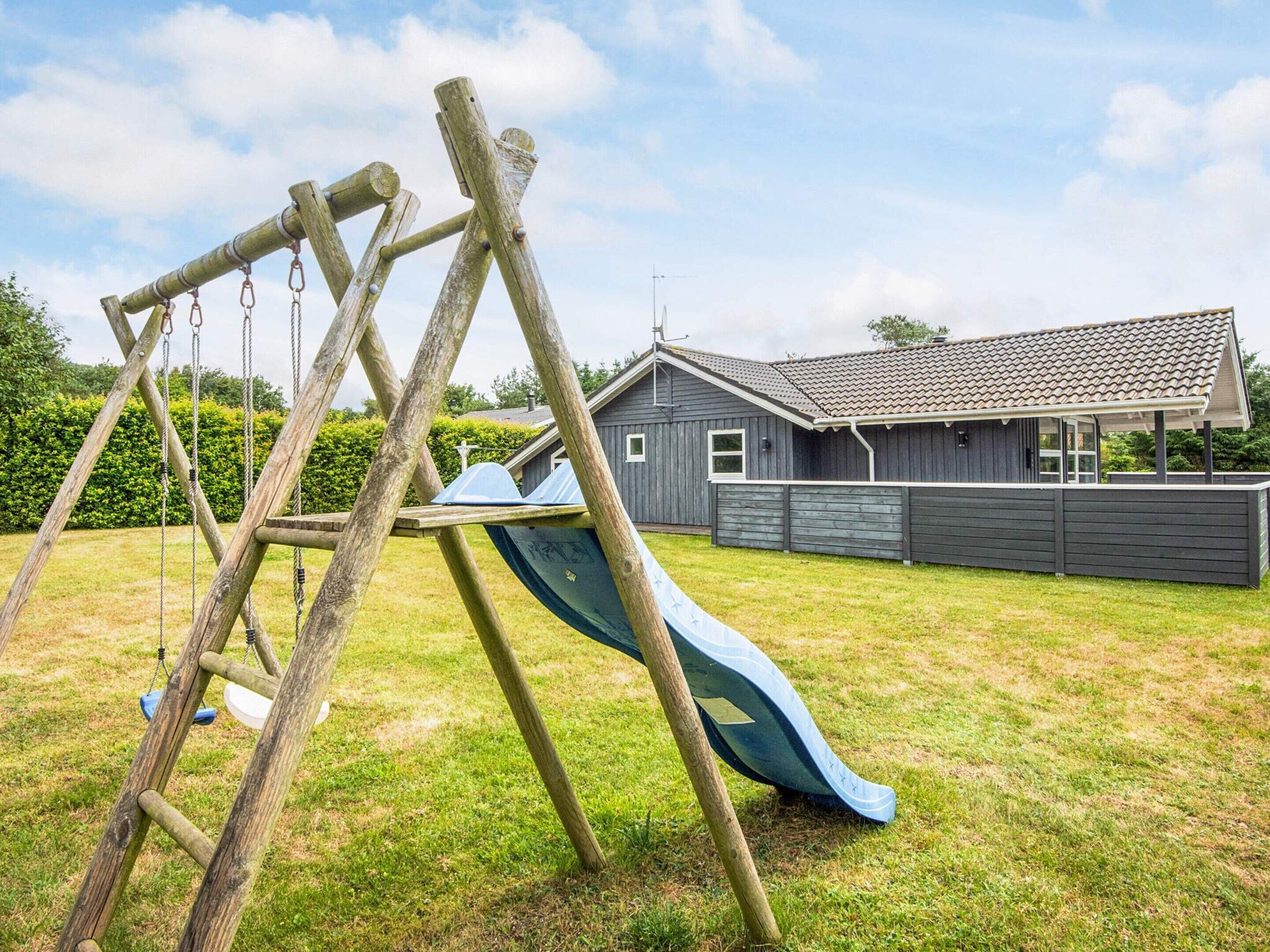 A wooden swing set and slide in a grassy yard with a house in the background.