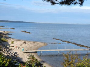 A beach with a wooden pier extending into calm water, surrounded by rocks and sparse vegetation.