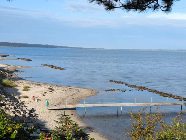 A beach with a wooden pier extending into calm water, surrounded by rocks and sparse vegetation.