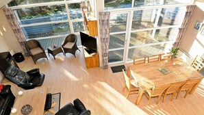 A spacious living area with a large wooden dining table, multiple chairs, and a sofa. There is a TV mounted on the wall and a window providing a view of the outdoors.