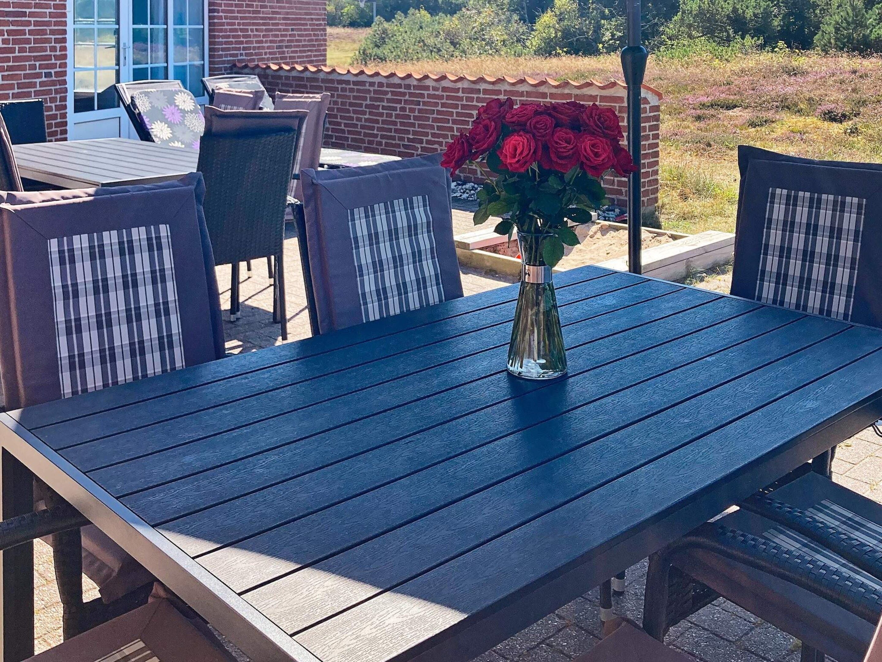 A patio with a table and chairs, a vase with red flowers, and a brick wall in the background.