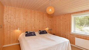 A bedroom with a wooden paneled wall, a bed with white bedding, two decorative pillows, a hanging light fixture, and a window with a view of trees.