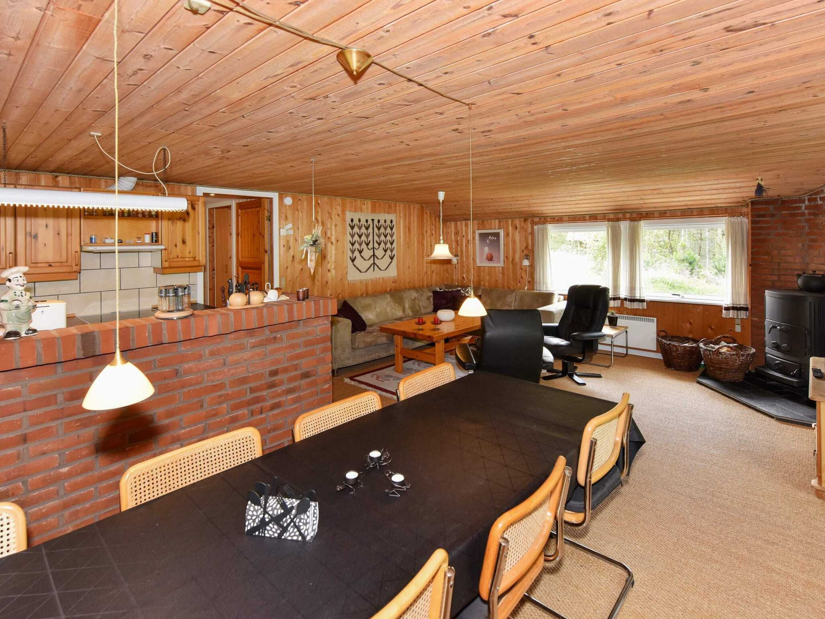 A cozy cabin interior with a wooden ceiling, a dining area with a black table and wicker chairs, a sofa, a fireplace, and a kitchen with wooden cabinets and a brick wall.