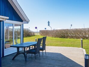 An outdoor deck with a table and chairs, a glass door leading to a room, and a view of a hedge and road signs.