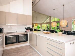 A modern kitchen with built-in appliances, a large island with seating, and large windows offering a view of greenery. - 5 Star Holiday Home in Henne (Henne)