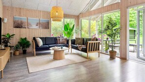 A spacious living area with a wooden floor, a black sofa, a coffee table, and large windows offering a view of greenery. - 5 Star Holiday Home in Henne (Henne)