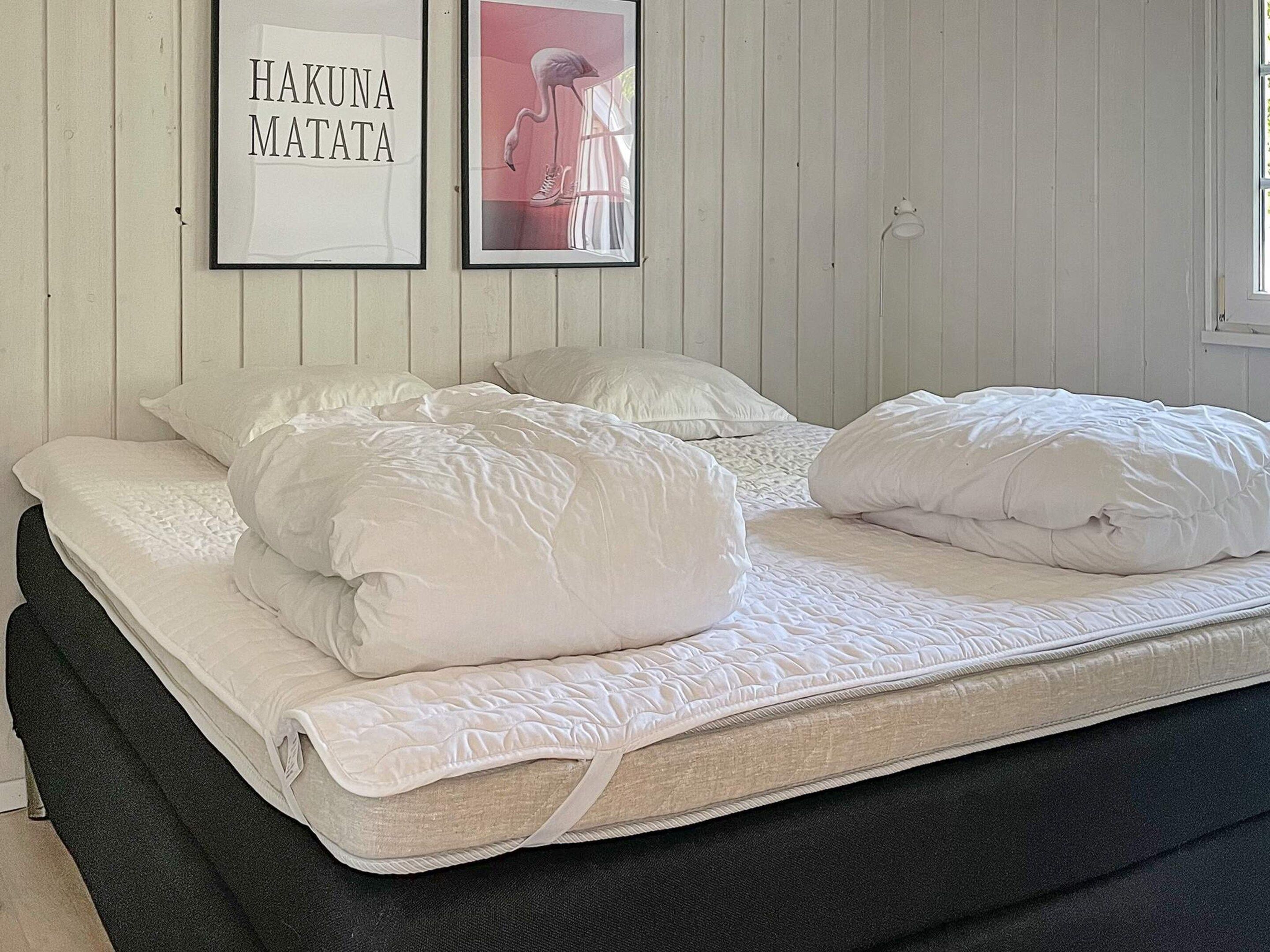 Una cama con ropa blanca y almohadas, un cuadro con texto y una lámpara en la pared.