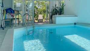 Piscina interna com água cristalina, uma escada e vista para área de estar externa e vegetação.