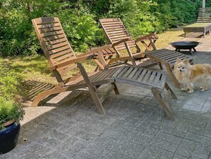Outdoor wooden lounge chairs arranged on a paved patio with a small round table and a black grill.
