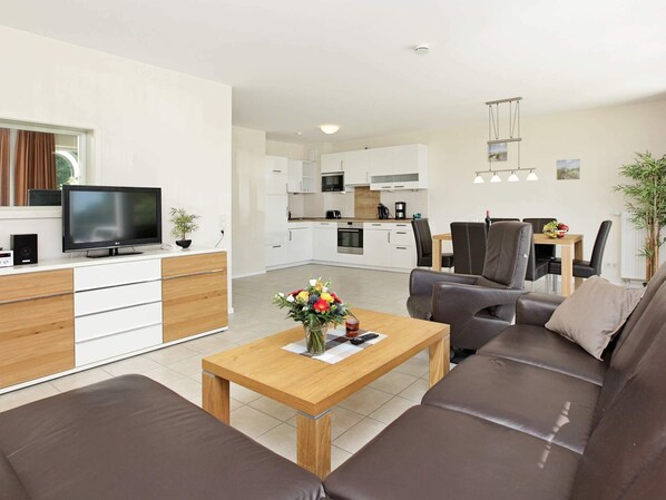 A modern living room with a sofa, a coffee table with flowers, a TV, and a kitchen in the background. - Holiday Home in Grossenbrode (Grossenbrode)