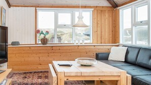 Une pièce chaleureuse avec un plafond en bois, un canapé, une table avec un bol et une télécommande, et une fenêtre donnant sur des maisons.